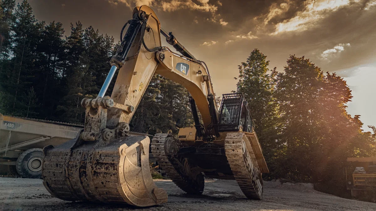 Großer CAT Kettenbagger mit abgesenktem Löffel, sichtbares Kettenlaufwerk, im Hintergrund Wald und Muldenkipper