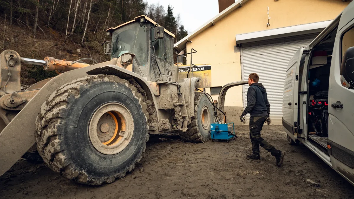 Mechaniker geht vom WIBAKO-Servicewagen zu einem verschlammten Radlader, an der Maschine ein blaues Servicegerät