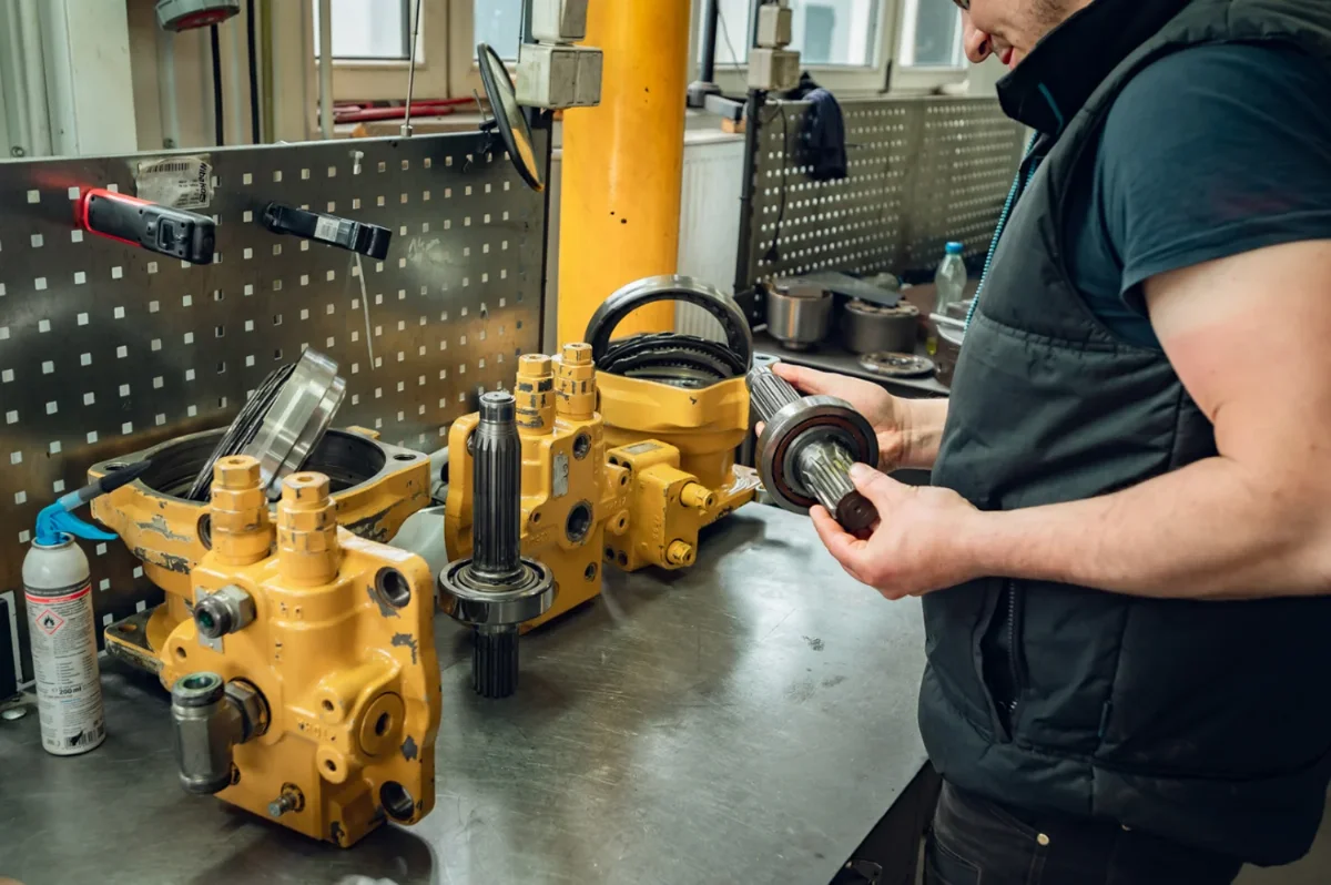 Mechaniker prüft eine Keilwelle mit Lager neben einem zerlegten gelben hydraulischen Schwenkmotor auf der Werkbank